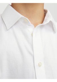 Gros plan sur le col d'une chemise blanche boutonnée et la partie haute de la poitrine avec deux boutons visibles, portée par une personne à la peau claire.
