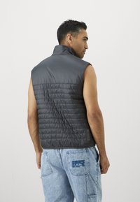 Sleeveless puffer vest in dark gray with a quilted texture, high collar, and smooth back; worn with light blue denim pants.