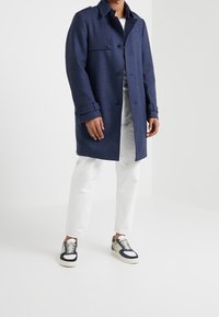 Cappotto in misto lana blu navy con chiusura a bottoni, maniche lunghe e tasche laterali, abbinato a pantaloni bianchi e scarpe sportive.