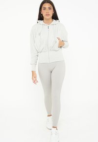 Felpa grigia chiara con zip, dotata di cordini e orlo a coste, abbinata a leggings a coste coordinati e sneakers bianche. Texture liscia in tutto il capo.