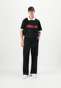 Black polo shirt with red graphic, white collar; paired with black cargo pants and white sneakers. Black cap completes the look.