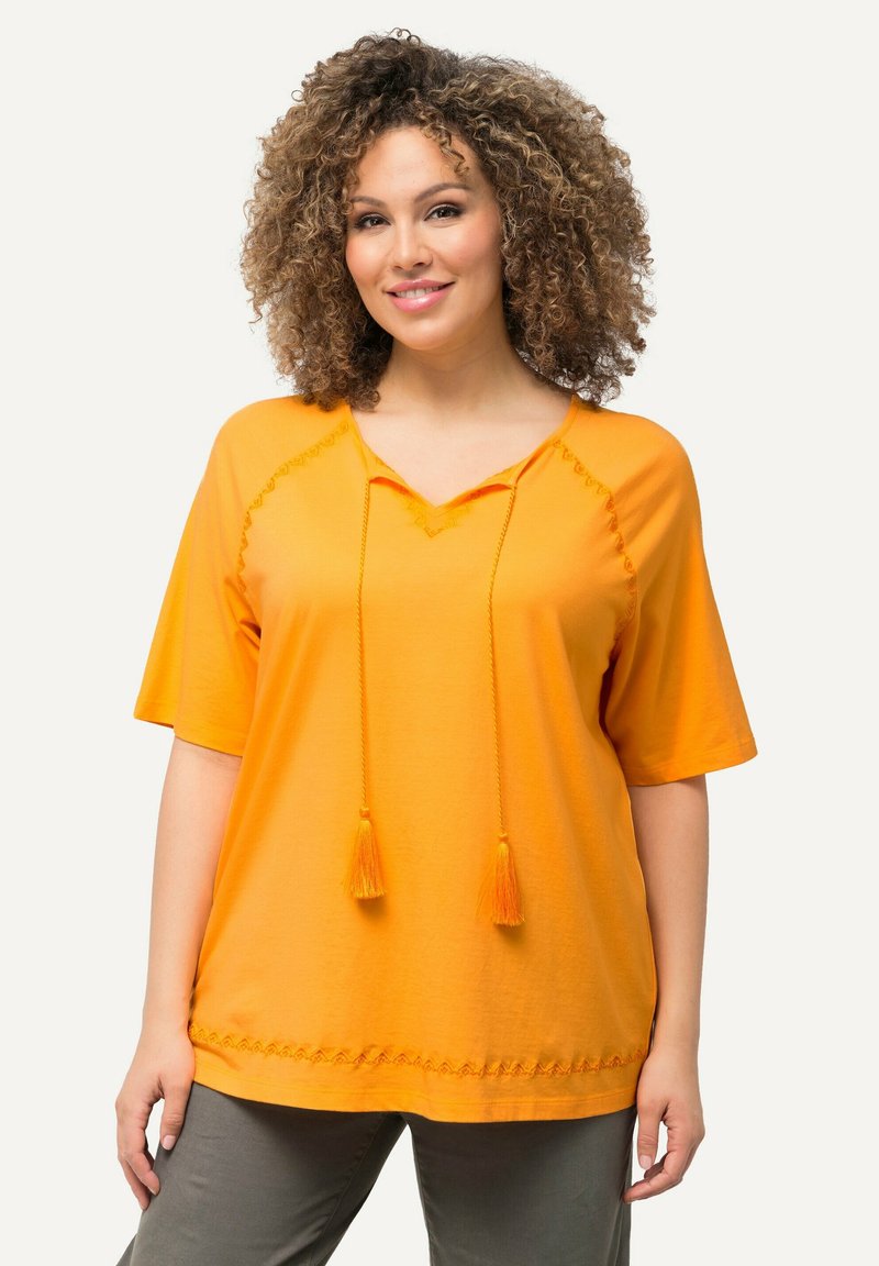 T-shirt in cotone arancione con maniche corte, scollatura a V, pon-pon decorativi e ricami lungo l'orlo. Vestibilità comoda.
