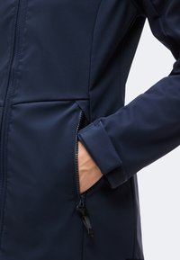 Main partiellement dans la poche zippée d'une veste bleu foncé avec une fermeture de poignet en velcro.