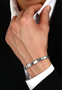 Silverfärgad metallarmband med platta länkar, slät yta, liten blå accentdetalj och ett säkert spänne. Bärs på ett handled med en vit skjorta.