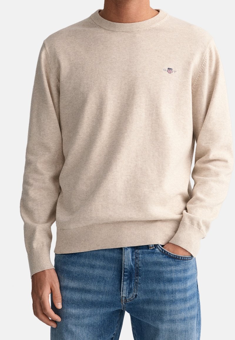 Pull beige en maille avec encolure ronde et poignets côtelés. Présente un petit logo brodé sur la poitrine. Associé à un jean bleu.