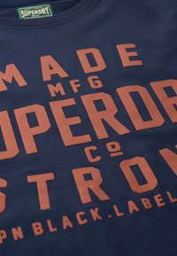 Camiseta de algodón azul marino con texto grande y en relieve en naranja que dice "SUPER" y "STRONG". Diseño de cuello redondo con etiqueta visible.