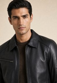 Mann mit dunklen Haaren, der eine schwarze Lederjacke über einem braunen Strickpullover trägt und direkt in die Kamera schaut vor einem einfarbigen Hintergrund.