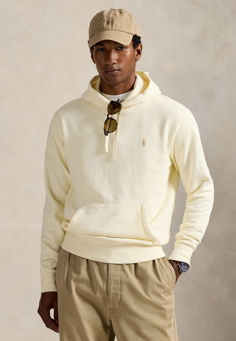 Uomo con cappellino beige, felpa crema con cappuccio e occhiali da sole appesi al cordoncino, pantaloni kaki e orologio da polso, in piedi davanti a uno sfondo semplice.