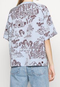 Kortärmad, ljusblå skjorta med ett brunt tropiskt landskapsmönster som inkluderar byggnader, palmer och fåglar i en avslappnad passform.