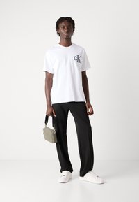 Hvid bomulds T-shirt med marineblå logo, sorte bukser, hvide sneakers og en lille grå crossbody taske. Enkel design, minimale detaljer.