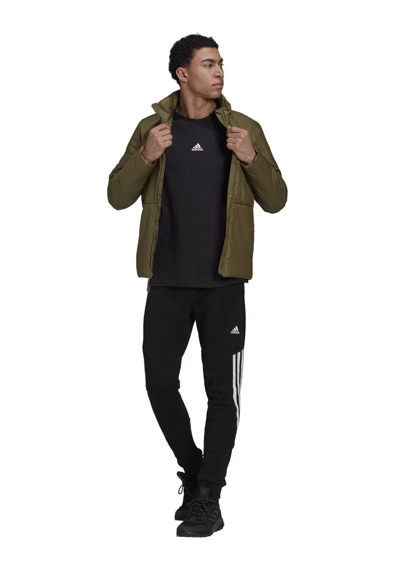 Giacca isolante verde oliva con zip nere, indossata sopra una t-shirt nera, abbinata a pantaloni tuta neri con strisce bianche. Calzature nere.