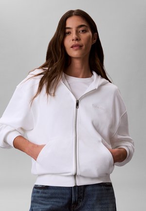 Pikka pruuni juustega naine, kes kannab valget lukuga kapuutsi ja tal on käed taskutes ning sinised teksad, seisab halli ühevärvilise tausta ees.