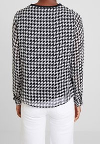 Blusa trasparente a righe bianche e nere a motivo chevron con maniche lunghe, scollatura rotonda e polsini arricciati, abbinata a pantaloni bianchi.