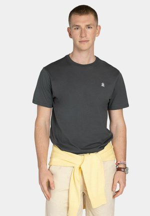 Joven vestido con una camiseta gris oscuro, pantalones beige y un suéter amarillo atado a la cintura, de pie frente a un fondo claro y sin adornos.