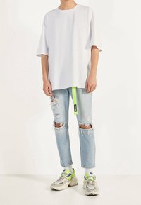 Person som bär en lös vit T-shirt, ljusa slitna jeans med färgfläckar, ett neongrönt bälte och vita sneakers med neonkant.