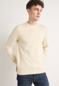 Light beige sweater with a round neckline, long sleeves, ribbed cuffs and hem, featuring a small textured logo detail on the front.