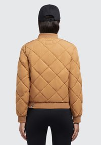 Gequilte, karamelkleurige pufferjack met een geribde zoom en manchetten. Heeft een hoge kraag en een klein label aan de achterkant. Soepele textuur.