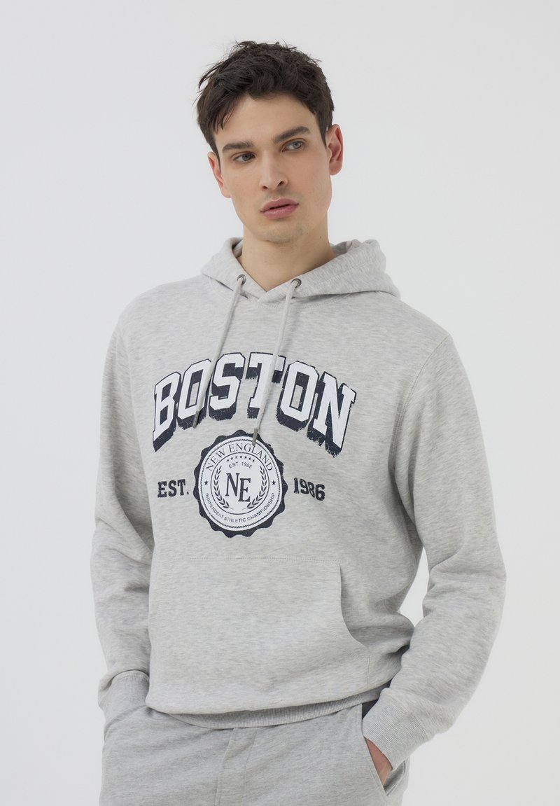 Felpa grigia con cappuccio regolabile e tasca frontale. Presenta la scritta "BOSTON" in grassetto blu navy e un logo grafico rotondo al centro. Tessuto morbido.