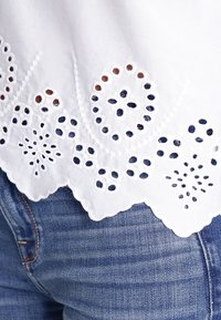 White embroidered blouse with scalloped hem, featuring eyelet detailing and floral patterns, paired with blue denim jeans.