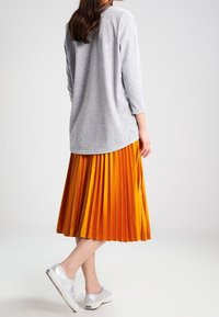 Grå långärmad topp, plisserad orange kjol och silverfärgade sneakers. Tyget har en mjuk textur; kjolen har vertikala plisseringar och varierande nyanser.