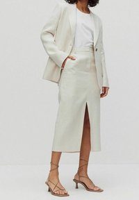 Světlý béžový přiléhavý blejzr přes bílou blůzu, doplněný odpovídající sukní ke kolenům s předním rozparkem a béžovými sandály na podpatku.