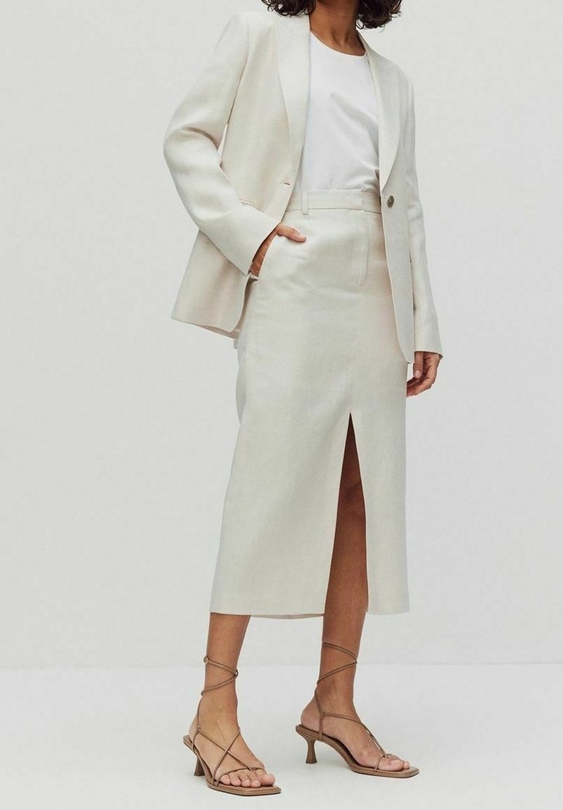 Světlý béžový přiléhavý blejzr přes bílou blůzu, doplněný odpovídající sukní ke kolenům s předním rozparkem a béžovými sandály na podpatku.