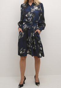 Een jurk van donkerblauwe stof met een schilderachtige zwart-wit patroon, knopen aan de voorkant, een iets versmalde taille, lange mouwen en zakken.