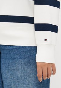 Hvit langermet genser med marineblå horisontale striper, med ribbestrikket krage og mansjetter, samt en liten logo nær mansjetten. Sammen med blå denim.