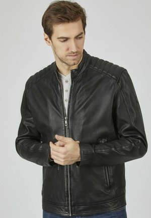 Chaqueta de cuero negra con detalles acanalados en los hombros, cierre de cremallera, collar tipo stand y dos bolsillos laterales. Textura suave, diseño ajustado.
