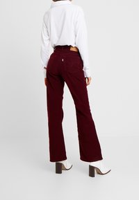 Pantalon évasé en velours côtelé bordeaux taille haute, poches arrière et patch en cuir de la marque au-dessus de la poche droite. Stylé avec une chemise blanche et des bottes.