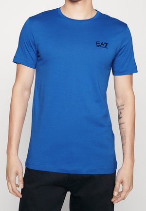 Hombre con camiseta azul lisa de EA7 Emporio Armani de manga corta y pantalones negros, mostrando tatuajes en el antebrazo derecho.