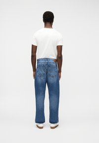 Uomo in piedi di spalle, indossa una camicia bianca a maniche corte, jeans larghi blu e sneakers bianche su uno sfondo chiaro e uniforme.