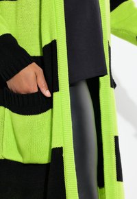 Cardigan lavorato a maglia con strisce verde neon e nere, con apertura frontale, polsini a costine e tasche frontali. Sotto si vedono un top nero e leggings lucidi.