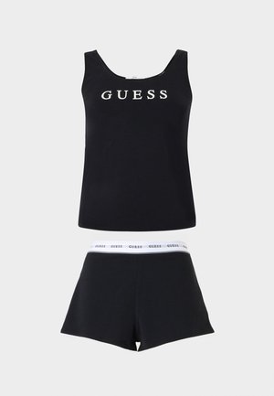 Juodas be rankovių viršus su baltais užrašu „GUESS“, derinamas su juodais šortais, kuriuose yra baltas juosmuo su nuolatiniu „GUESS“ spaudiniu.