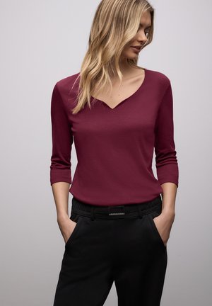 Bordowa bluzka z długim rękawem i dekoltem w kształcie litery V, wykonana z miękkiego materiału. Do kompletu czarne spodnie z elastycznym pasem i kieszeniami.
