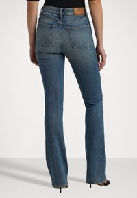 Jeans de mezclilla acampanados en un tono azul medio, con una cintura alta, cinco bolsillos y un parche de cuero en la parte trasera. Textura desvanecida en las piernas.