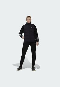 Zwarte puffervesting met hoge kraag over een zwart langemouwshirt, met witte gestreepte accenten, gecombineerd met zwarte leggings en sportschoenen.