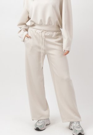 Osoba ubrana w beżowe szerokie spodnie dresowe ze ściągaczem w talii, dopasowaną, oversize bluzę i szare buty sportowe, stojąca z rękami w kieszeniach.