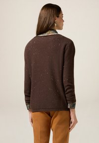 Brauner Pullover mit strukturierter Oberfläche und leichtem Sprenkel, mit einem gemusterten Kragen, der darunter hervorsieht, getragen über hellbraunen Hosen.