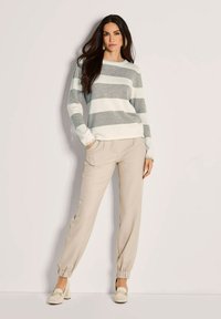 Frau steht mit einem grau-weißen gestreiften Pullover, beigen Jogginghosen und cremefarbenen Absatzschuhen vor einem einfarbigen Hintergrund.