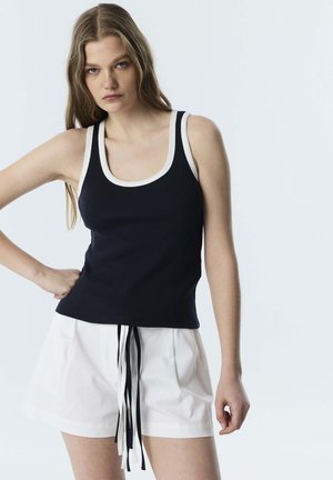 Jonge vrouw met lang haar die een zwart tanktopje met witte rand en witte korte broek met zwart-witte koordjes draagt tegen een lichte achtergrond.