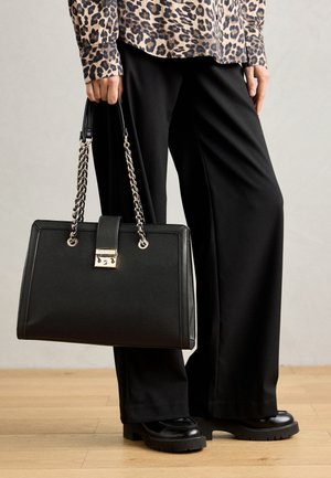 Personne tenant un sac à main en cuir noir avec des anses en chaîne, portant un pantalon noir à jambes larges, des chaussures noires et un top imprimé léopard.