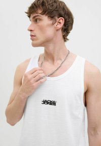 Bílý bezrukávový top vyrobený z lehkého materiálu, s malým černým logem na hrudi a stříbrným náhrdelníkem.