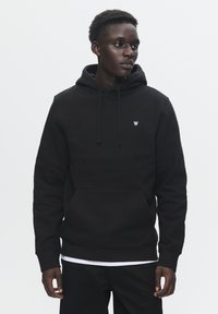 Black hooded sweatshirt made of textured fabric, featuring a front kangaroo pocket and subtle logo on the chest. Standard fit with drawstrings.