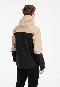 Vedenkestävä takki, jossa on beige ja musta väri-ero, säädettävä huppu ja sileä rakenne. Takakatsaus osoittaa rentoa istuvuutta.