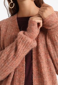 Texturovaný, žebrovaný cardigan v teplém broskvovém odstínu, vyrobený z chlupatého pleteného materiálu, má dlouhé rukávy a otevřený přední design.
