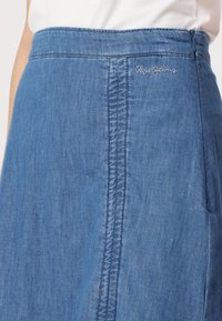 Denim krilo v srednje modri barvi, z gladko teksturo, navpičnim šivanjem ob strani in diskretno vezeno logotipom blizu pasu.
