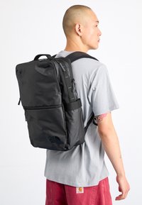 Svart nylon ryggsäck med rektangulär form, som har dragkedjefickor, justerbara remmar och en logotypdetalj på den nedre framsidan.