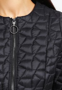 Chaqueta acolchada negra con costuras geométricas y un zipper plateado central con un tirador circular, llevada por una persona.