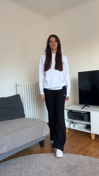 Sweatshirt branca com capuz e calças de treino pretas de corte largo, combinadas com ténis brancos; em pé num chão de madeira.